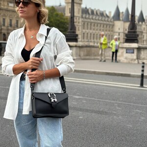 Puede incluir: Una mujer con una camisa de lino blanca, pantalones vaqueros azules y gafas de sol lleva un bolso bandolera de cuero negro con una hebilla plateada. El bolso tiene una correa larga y se lleva sobre el hombro.