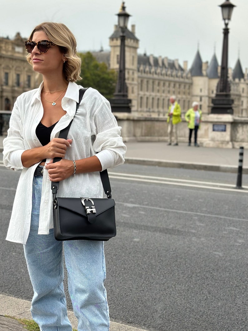 Puede incluir: Una mujer con una camisa de lino blanca, pantalones vaqueros azules y gafas de sol lleva un bolso bandolera de cuero negro con una hebilla plateada. El bolso tiene una correa larga y se lleva sobre el hombro.