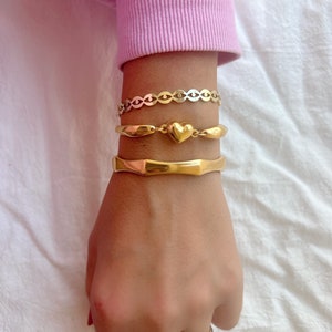 May include: A set of three gold bracelets on a wrist. The top bracelet is a delicate chain with a heart charm. The middle bracelet is a thick, textured band. The bottom bracelet is a simple, smooth band.