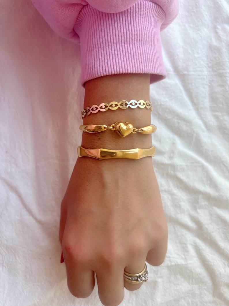 May include: A set of three gold bracelets on a wrist. The top bracelet is a delicate chain with a heart charm. The middle bracelet is a thick, textured band. The bottom bracelet is a simple, smooth band.