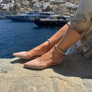 May include: A pair of light pink leather ankle strap sandals with a pointed toe. The sandals are worn by a person sitting on a rocky surface with a view of a town and boats in the distance.