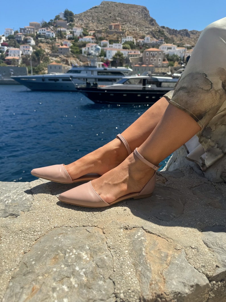 May include: A pair of light pink leather ankle strap sandals with a pointed toe. The sandals are worn by a person sitting on a rocky surface with a view of a town and boats in the distance.