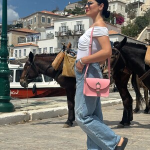 Op de afbeelding: Een vrouw in een wit off-shoulder topje, blauwe jeans en zwarte sandalen loopt over straat. Ze draagt een roze crossbody tas met een gouden gesp. Er zijn twee ezels op de achtergrond.