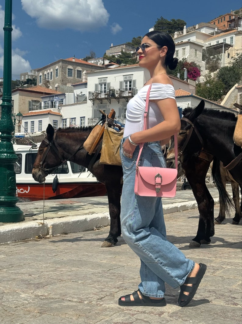 Op de afbeelding: Een vrouw in een wit off-shoulder topje, blauwe jeans en zwarte sandalen loopt over straat. Ze draagt een roze crossbody tas met een gouden gesp. Er zijn twee ezels op de achtergrond.