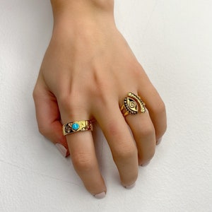 May include: Two gold rings on a hand. One ring has a turquoise stone and the other ring has an eye design.