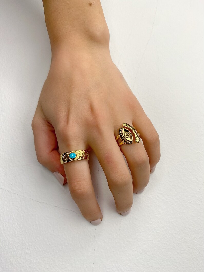 May include: Two gold rings on a hand. One ring has a turquoise stone and the other ring has an eye design.
