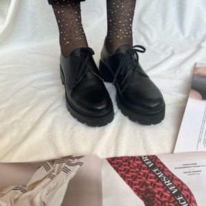 May include: Black leather lace-up shoes with thick soles and a chunky heel. The shoes are worn with black sheer socks with silver glitter.