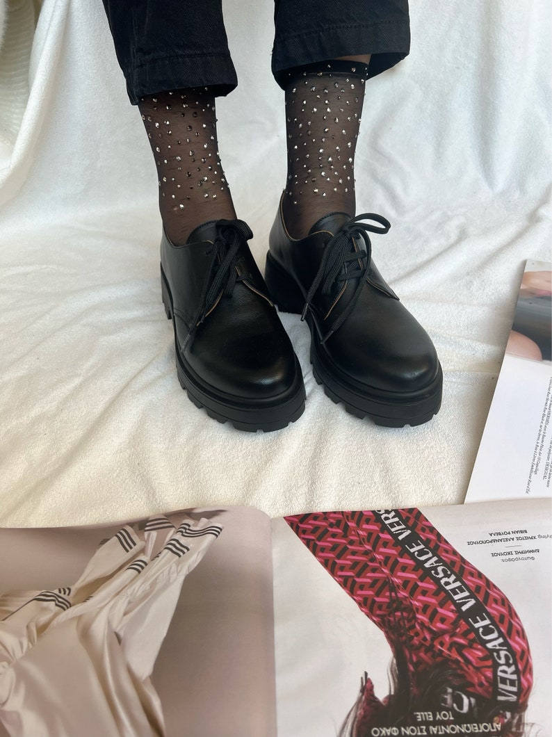 May include: Black leather lace-up shoes with thick soles and a chunky heel. The shoes are worn with black sheer socks with silver glitter.