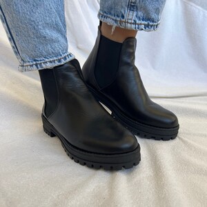 Pu&ograve; includere: Stivali Chelsea in pelle nera con suola spessa. Gli stivali hanno pannelli laterali elastici e un tirante sul retro. Indossati con jeans blu.