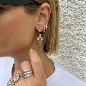 May include: Silver hoop earrings with a silver eye charm pendant. The earrings are worn in the earlobe and the charm is hanging down. The person is also wearing several silver rings on their fingers.