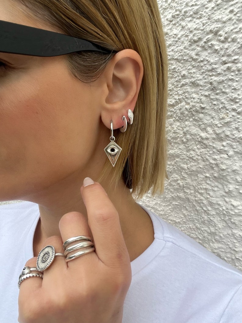 May include: Silver hoop earrings with a silver eye charm pendant. The earrings are worn in the earlobe and the charm is hanging down. The person is also wearing several silver rings on their fingers.