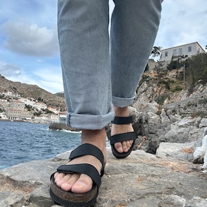 May include: Black sandals with two adjustable straps and a cork-style sole. The sandals are being worn outdoors, with the ocean and a coastal town in the background. The person is wearing light blue jeans.