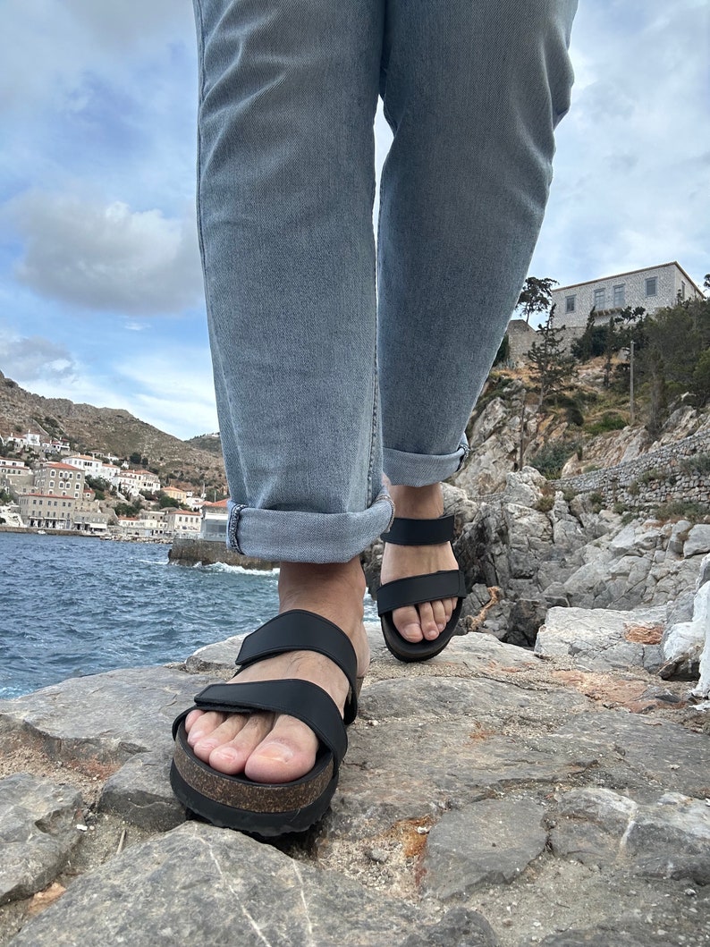 May include: Black sandals with two adjustable straps and a cork-style sole. The sandals are being worn outdoors, with the ocean and a coastal town in the background. The person is wearing light blue jeans.