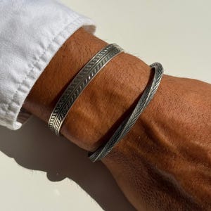 Può includere: Due bracciali a fascia in metallo color argento indossati al polso. Il primo bracciale presenta un motivo a linee verticali centrali con bordi lisci. Il secondo bracciale mostra un motivo dettagliato e testurizzato a spina di pesce sulla superficie.