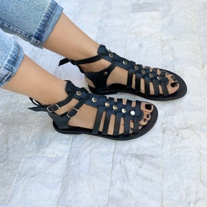 Puede incluir: Sandalias gladiadoras de cuero negro con m&uacute;ltiples correas y tachuelas plateadas. Las sandalias tienen una suela plana y una correa de tobillo con cierre de hebilla.