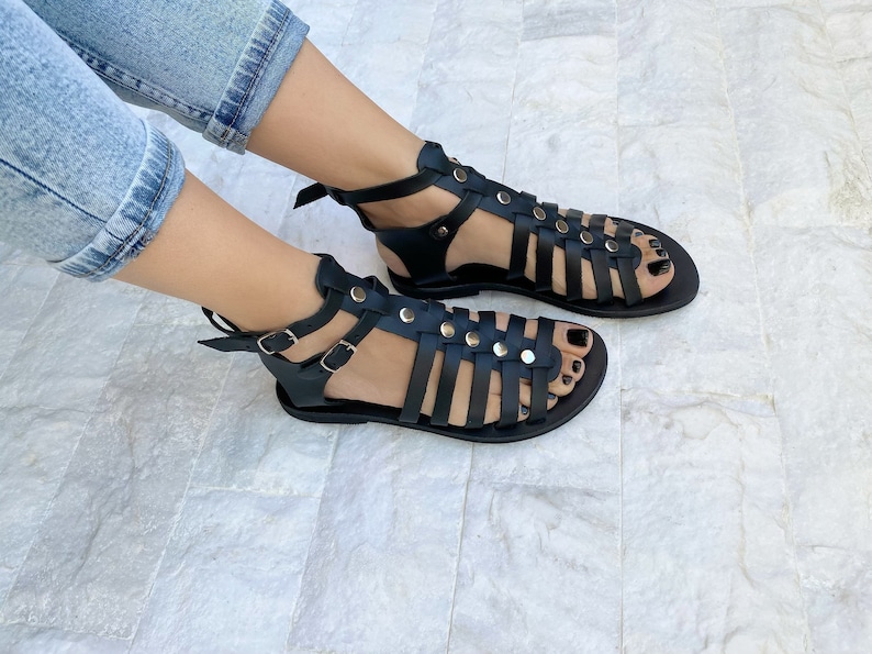 Puede incluir: Sandalias gladiadoras de cuero negro con m&uacute;ltiples correas y tachuelas plateadas. Las sandalias tienen una suela plana y una correa de tobillo con cierre de hebilla.