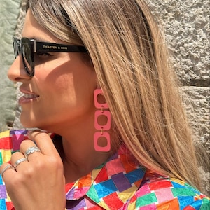 May include: A woman wearing pink square-shaped earrings with gold accents. She is wearing a colorful patterned shirt and black sunglasses with the text "KAPTEN & SON" on the frame.