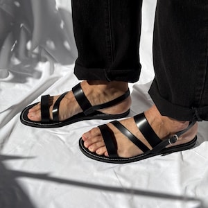 K&ouml;nnte beinhalten: Schwarze Ledersandalen mit mehreren Riemen und einem Schnallenverschluss. Die Sandalen haben ein minimalistisches Design mit wei&szlig;en N&auml;hten um die Sohle. Getragen mit einer schwarzen Hose.
