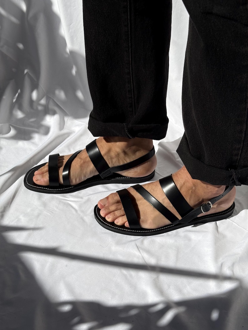 K&ouml;nnte beinhalten: Schwarze Ledersandalen mit mehreren Riemen und einem Schnallenverschluss. Die Sandalen haben ein minimalistisches Design mit wei&szlig;en N&auml;hten um die Sohle. Getragen mit einer schwarzen Hose.