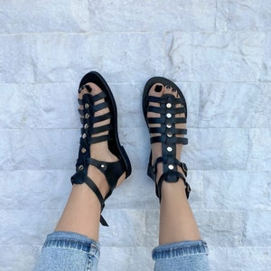 Puede incluir: Sandalias gladiadoras de cuero negro con hebillas y tachuelas doradas. Las sandalias tienen m&uacute;ltiples correas y se usan en los pies.