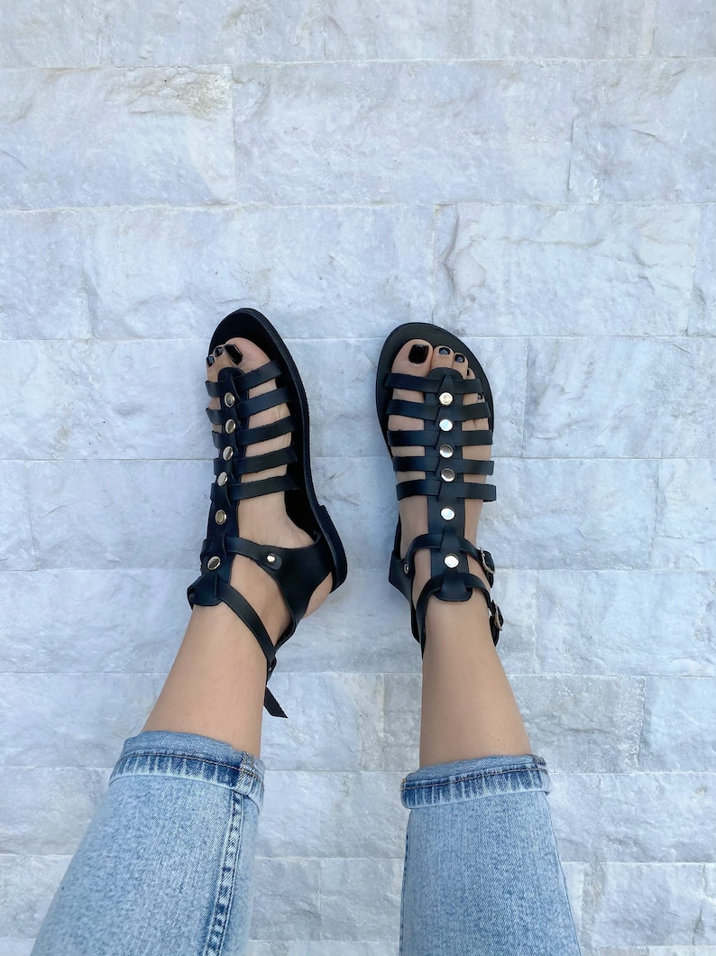 Puede incluir: Sandalias gladiadoras de cuero negro con hebillas y tachuelas doradas. Las sandalias tienen m&uacute;ltiples correas y se usan en los pies.