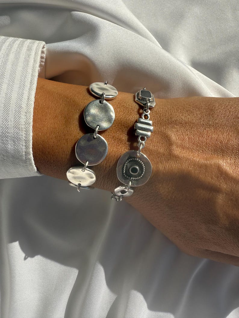 May include: Two silver bracelets with unique, textured designs. One bracelet features a series of connected, hammered-look circles and ovals. The other has a mix of geometric shapes and a circular pendant with a central design. The bracelets are displayed on a white background.