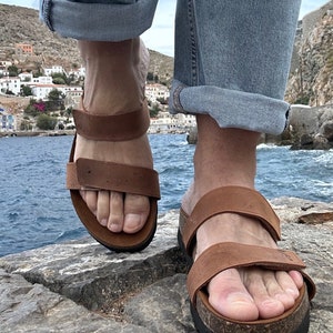 May include: Brown leather sandals with adjustable straps and a cork footbed. The sandals are worn with rolled-up blue jeans. The background shows a coastal town with white buildings and a blue sea.