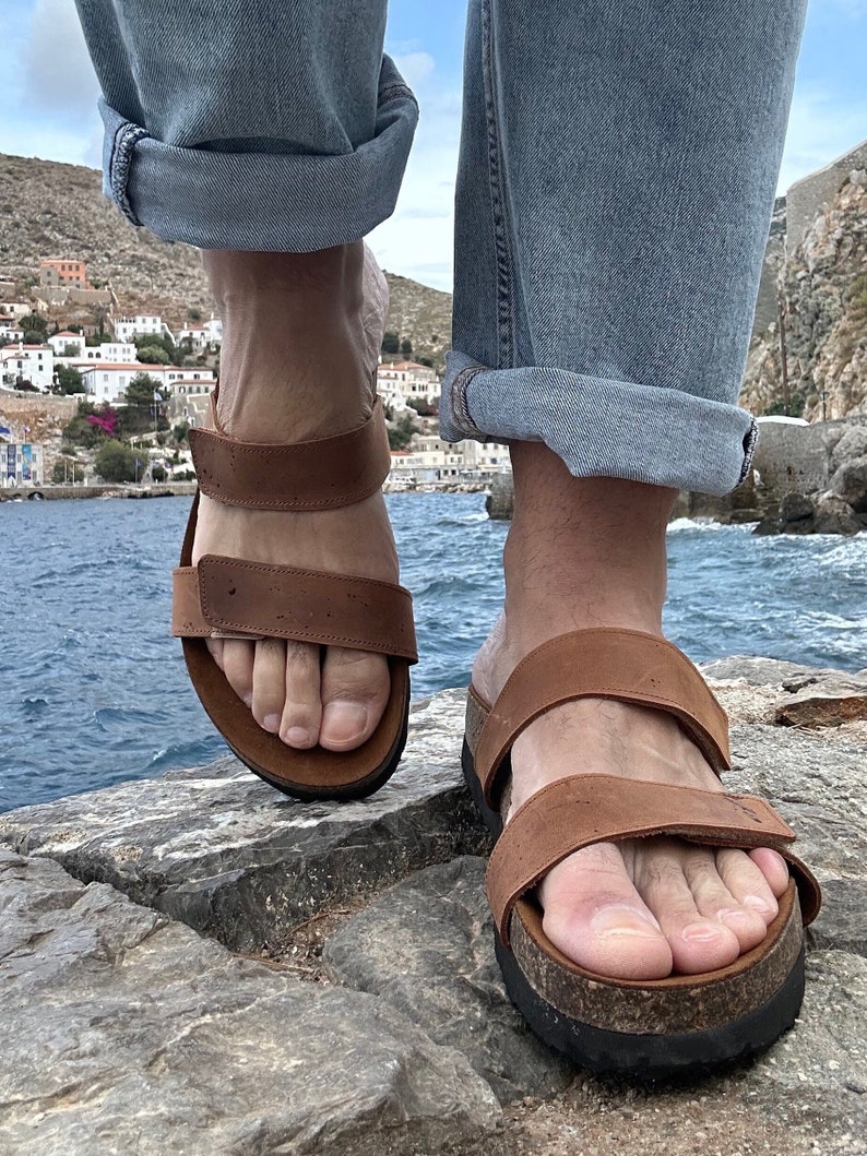 May include: Brown leather sandals with adjustable straps and a cork footbed. The sandals are worn with rolled-up blue jeans. The background shows a coastal town with white buildings and a blue sea.