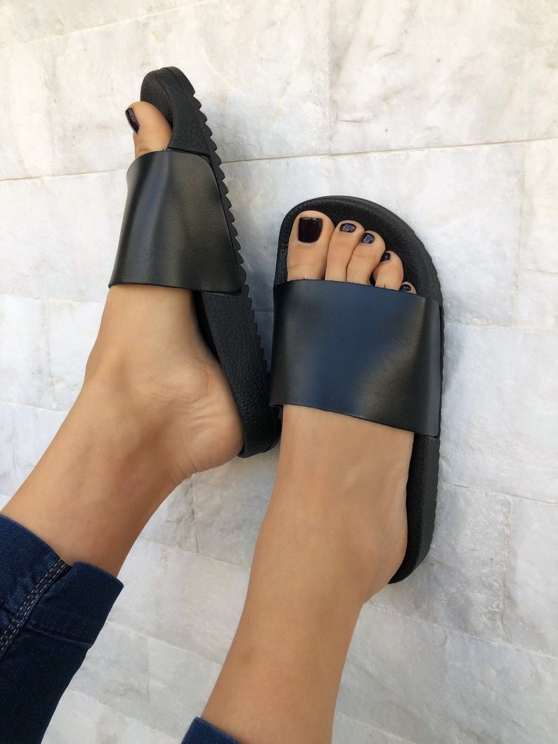 May include: Pair of black leather slide sandals with a textured rubber sole. The sandals have a simple design and are perfect for casual wear.