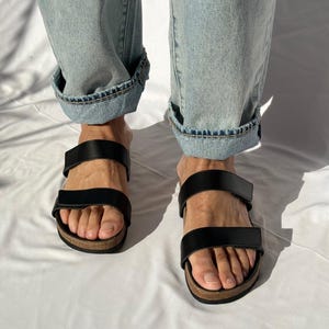 May include: Black leather sandals with two adjustable straps and a cork footbed. The sandals are worn with light blue denim jeans with cuffed hems. The sandals are a casual footwear choice.