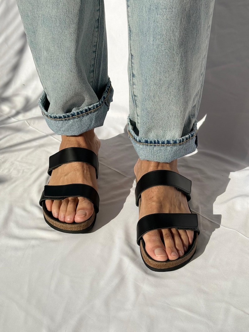May include: Black leather sandals with two adjustable straps and a cork footbed. The sandals are worn with light blue denim jeans with cuffed hems. The sandals are a casual footwear choice.