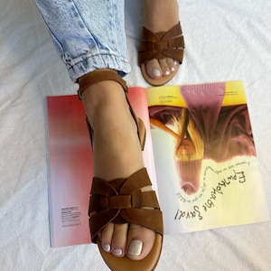 K&ouml;nnte beinhalten: Ein Paar braune Ledersandalen mit einem Kreuzriemen-Design. Die Sandalen haben einen Schnallenverschluss und einen niedrigen Absatz.