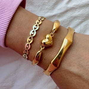 May include: Three gold bracelets on a wrist. The top bracelet is a delicate chain with a cutout pattern. The middle bracelet has a heart charm and is connected to a thicker, curved bracelet. The bottom bracelet is a thick, curved bangle.