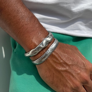 Może przedstawiać: Dwie srebrne bransoletki na czyimś nadgarstku. Obie bransoletki są fakturowane i mają młotkowane wykończenie. Górna bransoletka jest szersza i ma bardziej organiczny kształt. Dolna bransoletka jest cieńsza i ma bardziej geometryczny kształt.