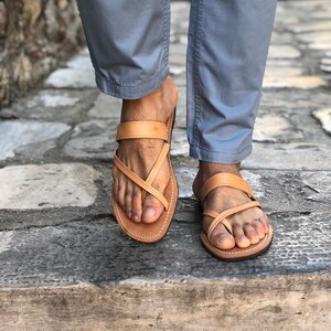 May include: A pair of tan leather sandals with criss-cross straps. The sandals are worn on a person's feet.