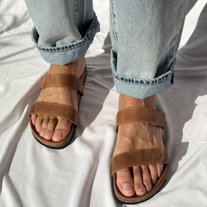May include: Brown leather sandals with two adjustable straps. The sandals have a cork footbed and are worn with light blue denim jeans with cuffed hems. The image is taken from a high angle.