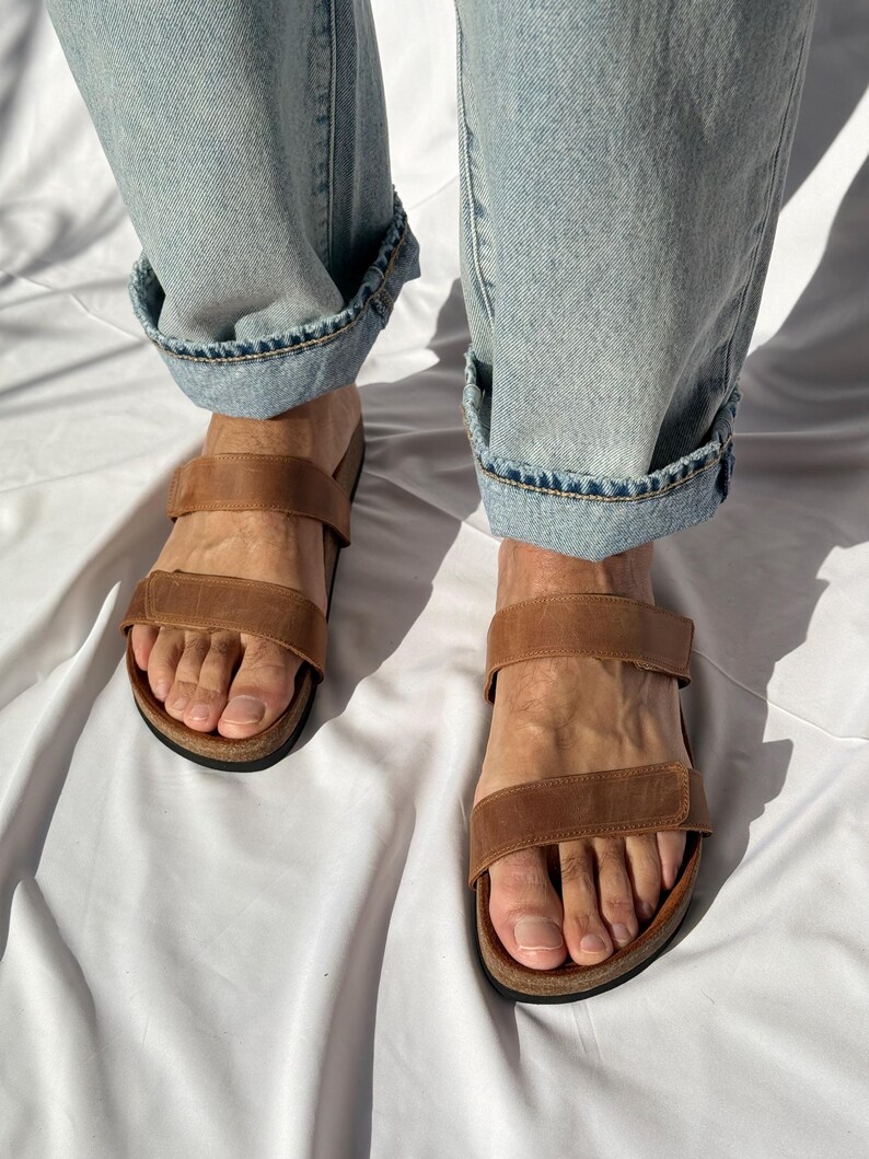 May include: Brown leather sandals with two adjustable straps. The sandals have a cork footbed and are worn with light blue denim jeans with cuffed hems. The image is taken from a high angle.
