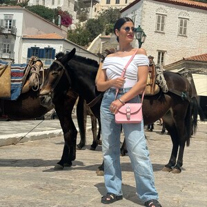 Op de afbeelding: Een vrouw in een wit off-shoulder topje, blauwe jeans en zwarte sandalen staat voor twee bruine ezels. Ze draagt een roze crossbody tas met een zilveren gesp. De ezels dragen zadels en hoofdstellen.