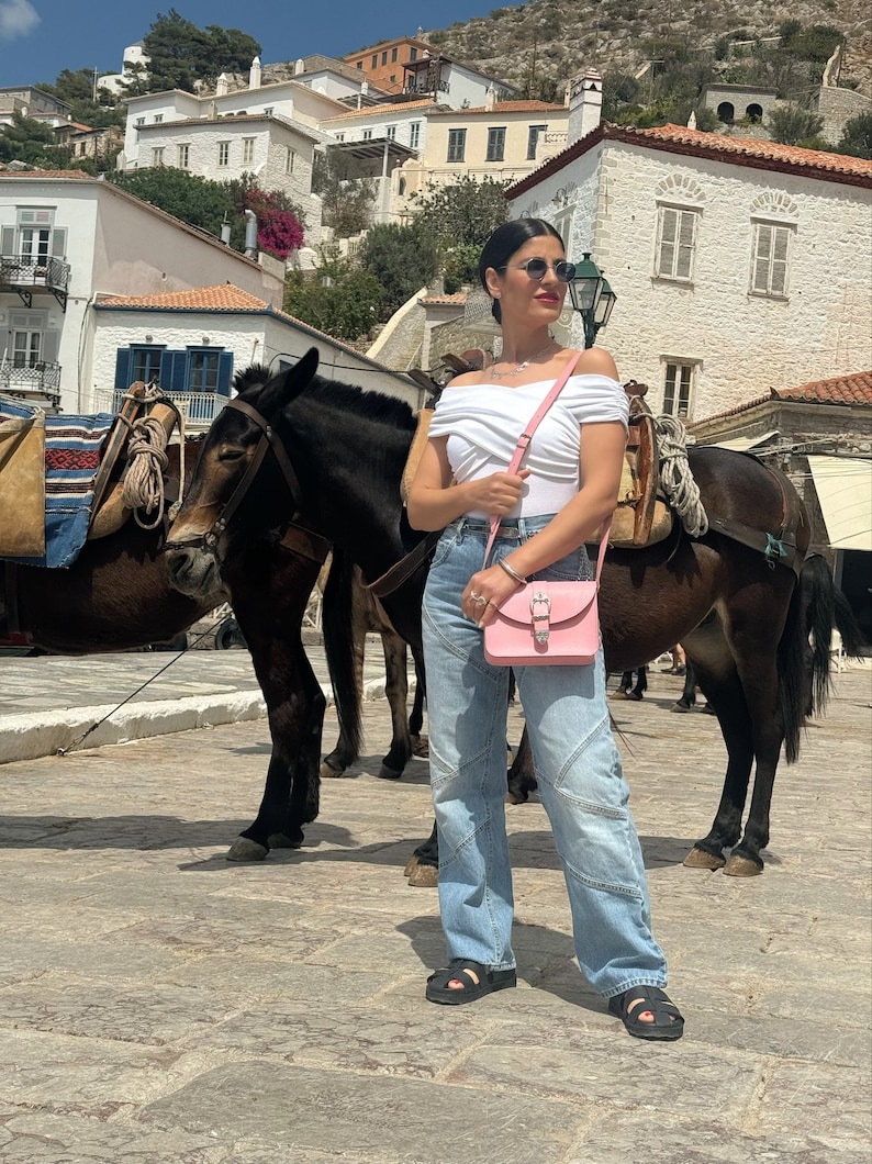 Op de afbeelding: Een vrouw in een wit off-shoulder topje, blauwe jeans en zwarte sandalen staat voor twee bruine ezels. Ze draagt een roze crossbody tas met een zilveren gesp. De ezels dragen zadels en hoofdstellen.