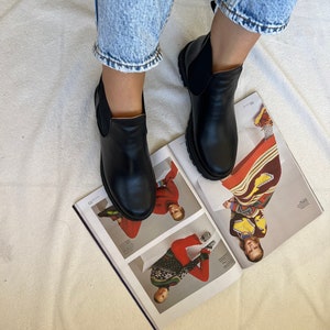 Pu&ograve; includere: Stivali Chelsea in pelle nera con pannelli laterali elastici. Gli stivali hanno la punta arrotondata e una suola spessa. Gli stivali sono indossati con jeans di denim blu chiaro. Gli stivali sono appoggiati su una rivista aperta.