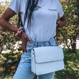 Puede incluir: Un bolso blanco de bandolera con cierre de solapa. El bolso est&aacute; hecho de cuero texturizado y tiene una correa de cadena plateada.