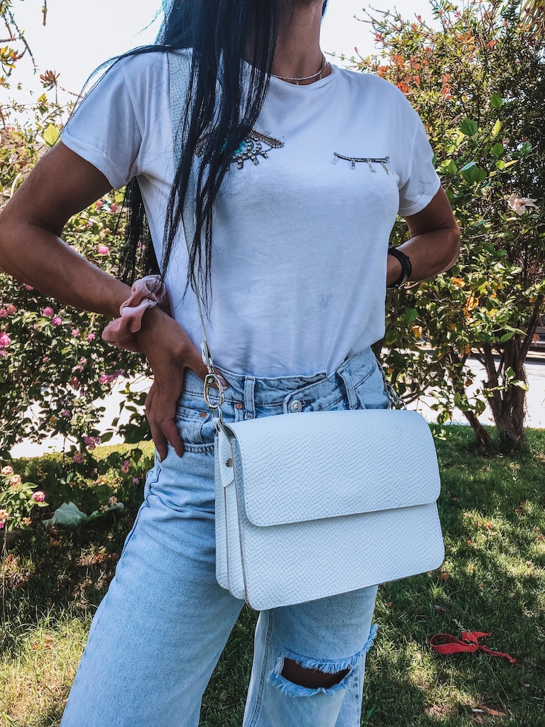 Puede incluir: Un bolso blanco de bandolera con cierre de solapa. El bolso est&aacute; hecho de cuero texturizado y tiene una correa de cadena plateada.