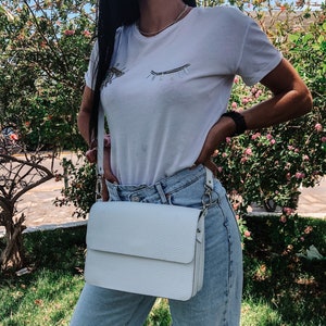 Puede incluir: Un bolso blanco de bandolera con cierre de solapa y una cadena plateada. El bolso lo lleva una persona que lleva una camiseta blanca con un dise&ntilde;o de ojo brillante y unos vaqueros azules claros.