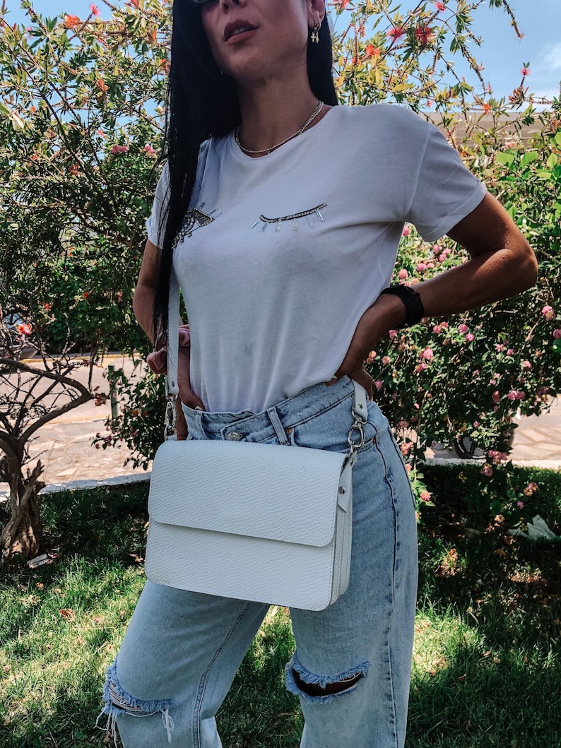 Puede incluir: Un bolso blanco de bandolera con cierre de solapa y una cadena plateada. El bolso lo lleva una persona que lleva una camiseta blanca con un dise&ntilde;o de ojo brillante y unos vaqueros azules claros.