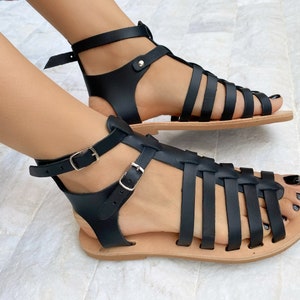 May include: Black gladiator sandals featuring multiple horizontal straps across the foot, a vertical center strap, and two adjustable ankle straps with silver-toned metal buckles. The sandals have a flat, light tan sole and an open-toe design.