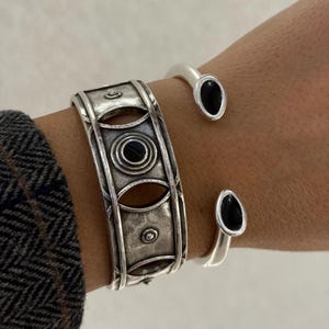 Könnte beinhalten: Zwei silberne Armbänder mit schwarzen ovalen Akzenten. Eines ist ein breites Armband mit kreisförmigen und ovalen Aussparungen, das andere ein schlankes offenes Armband. Die Armbänder werden an einem Handgelenk getragen.