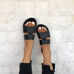 Puede incluir: Sandalias de cuero negro con un dise&ntilde;o minimalista. Las sandalias tienen una correa ancha en la parte superior del pie y otra correa cerca del tobillo. Combinadas con vaqueros negros.