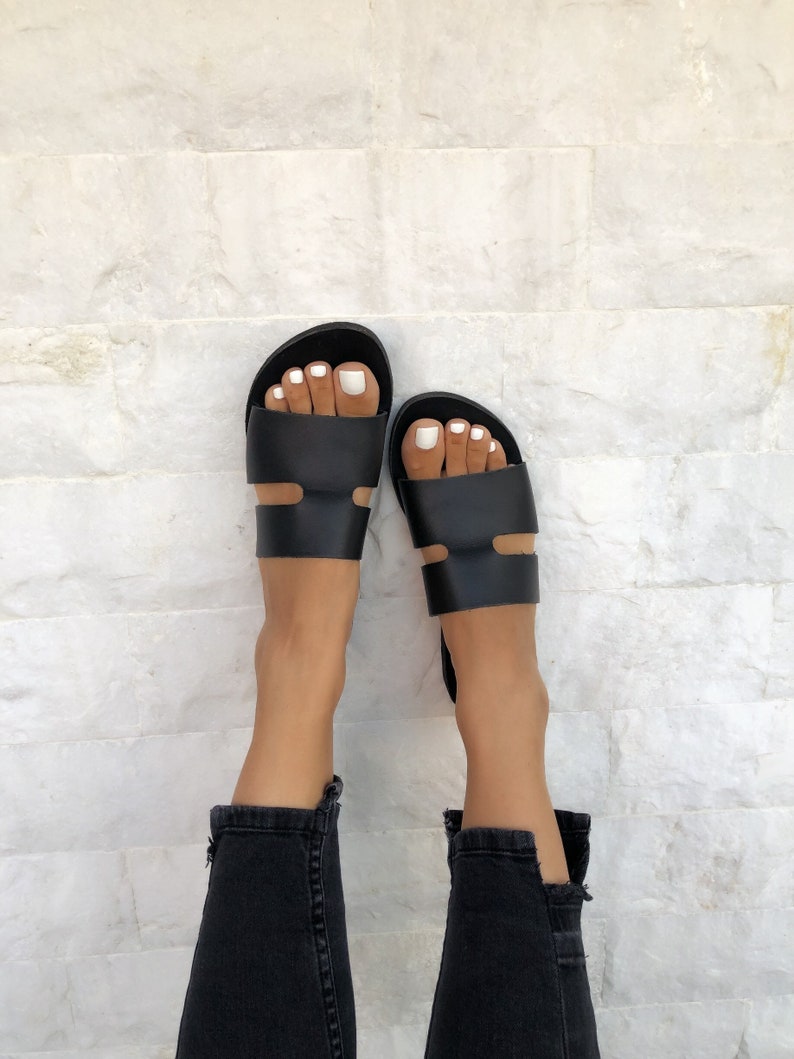 Puede incluir: Sandalias de cuero negro con un dise&ntilde;o minimalista. Las sandalias tienen una correa ancha en la parte superior del pie y otra correa cerca del tobillo. Combinadas con vaqueros negros.