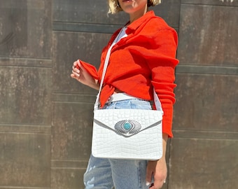 Può includere: Una borsa a tracolla bianca in pelle di coccodrilo con un ornamento in argento e turchese. La borsa è indossata da una persona che indossa una camicia rossa e jeans blu.