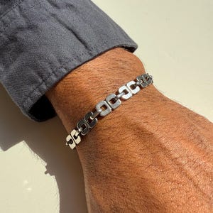 May include: A silver bracelet with a series of connected, rectangular links. The bracelet is worn on a wrist, with a gray shirt sleeve visible in the background. The bracelet's design is simple and modern.